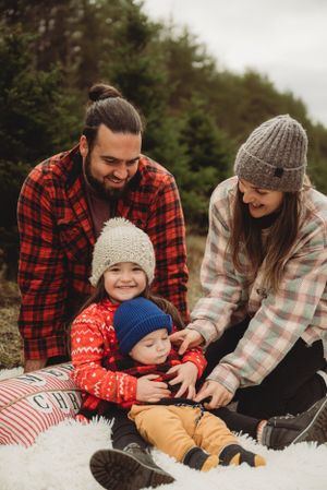 Family sitting together on blanket at Christmas tree farm wearing cozy winter clothing.