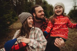 A joyful outdoor family portrait in winter clothing with knit hats against a wooded backdrop.