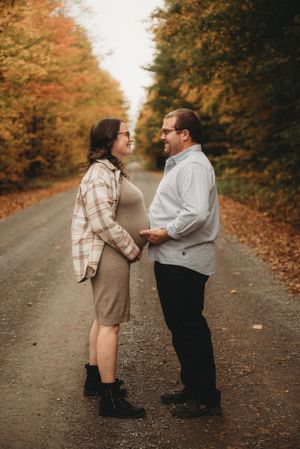Maternity couple embracing on autumn road lined with colorful fall foliage.