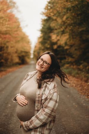 A pregnant woman wearing a plaid shirt smiles on a fall-lined road with autumn foliage in the background.