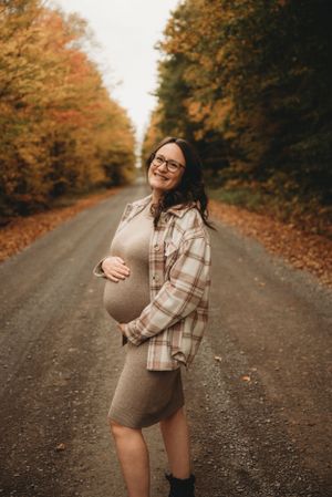 Expectant mother in plaid shirt smiling on tree-lined autumn road.