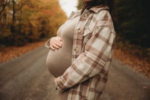 Close-up of pregnant belly in tan dress and plaid shirt on fall pathway.