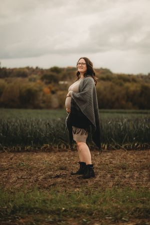 A maternity photo showing a pregnant woman in a gray cardigan standing in a field during fall.