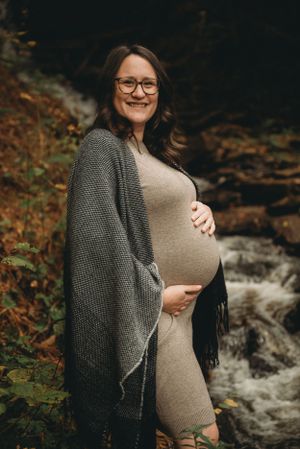 A maternity portrait by a waterfall featuring a pregnant woman in a knit cardigan and fitted dress.