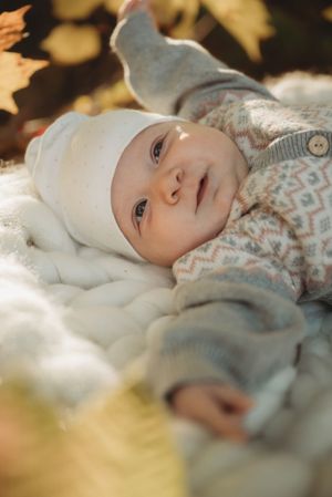 A baby wearing a white cap and patterned sweater rests on soft white blankets in natural light.