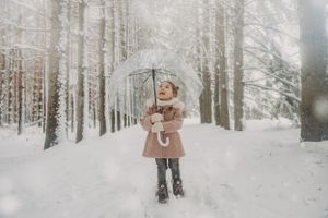 A joyful child plays in the snow among tall winter trees wearing a brown coat.