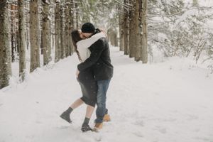 Couple embracing and lifting in joy on a snow-covered forest path.