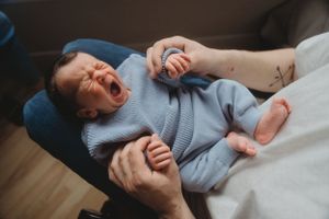Baby in blue sweater laughing while lying on white bedding.
