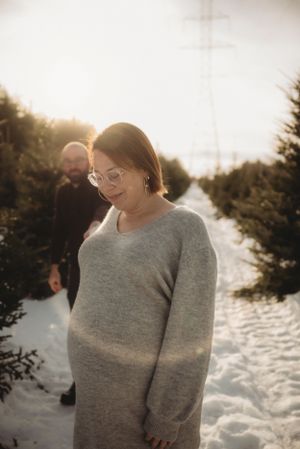 Person in gray sweater dress stands in snowy landscape at sunset among evergreen trees.