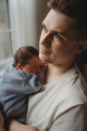 Parent holding newborn baby in blue knit sweater by window in natural light.
