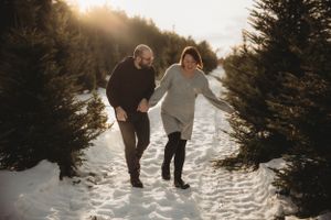 Couple walking together playfully through snowy path surrounded by evergreen trees at sunset.