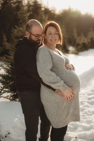 Couple shares tender moment in snowy winter setting with sunlight filtering through trees.