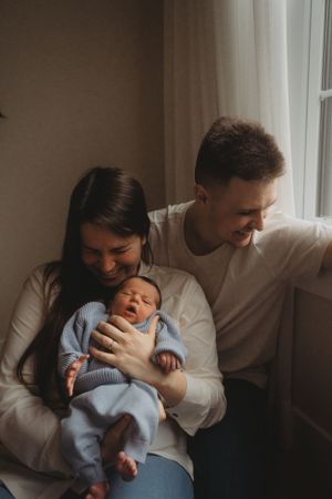 A joyful family moment by the window with a sleeping newborn in blue clothing.