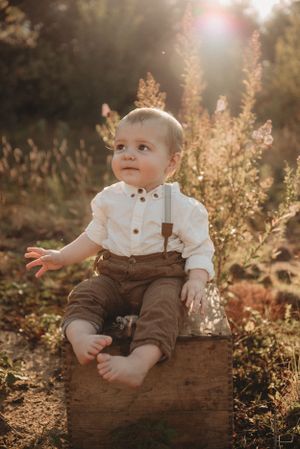 A small child sits on a wooden box outdoors wearing brown overalls and a white shirt.