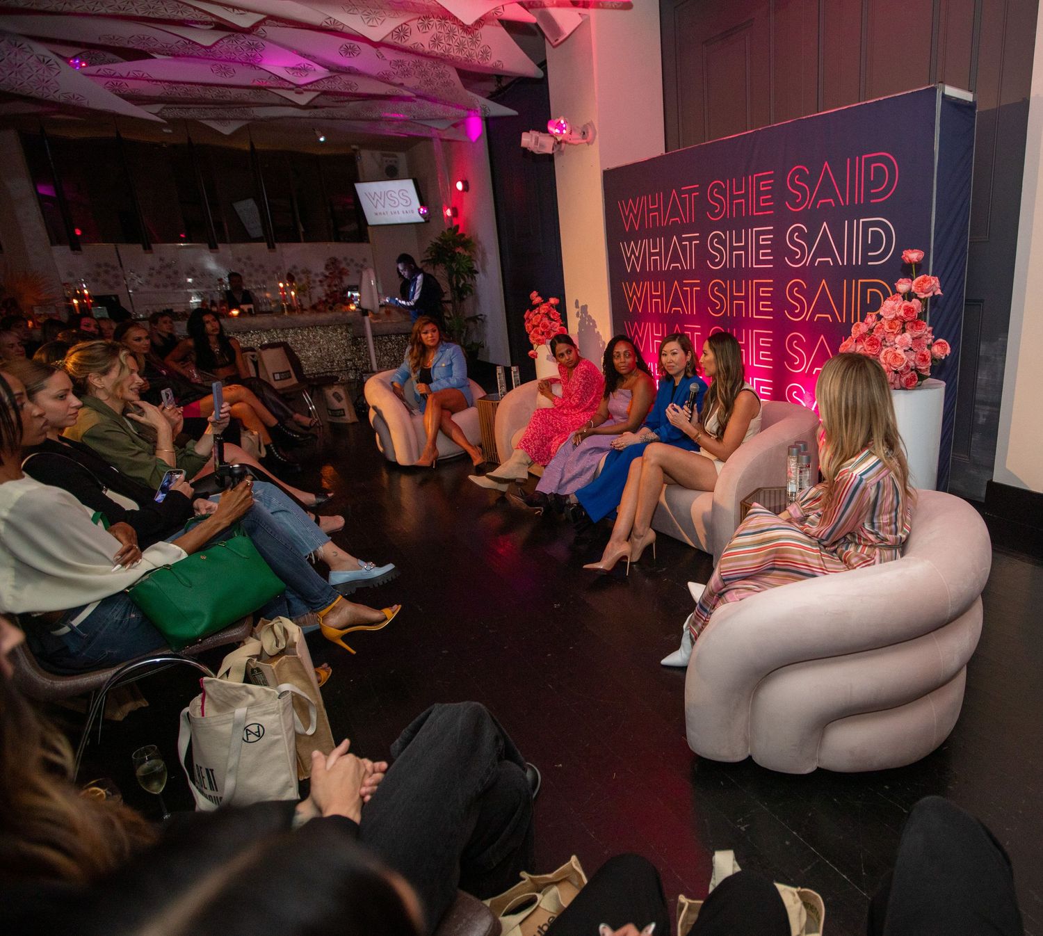Audience seated at an event with pink lighting and a neon sign display in the background.