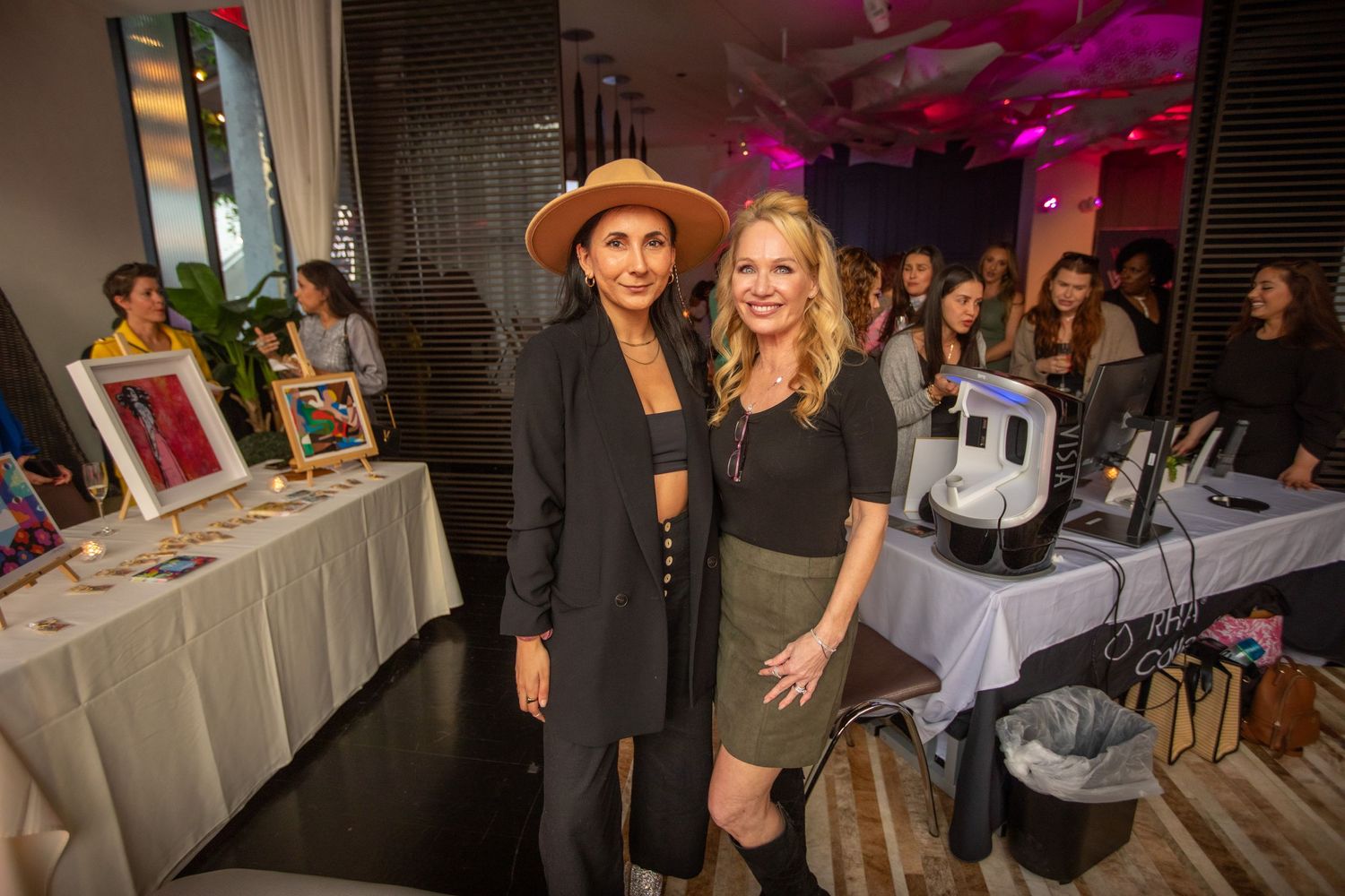 Vendors pose at beauty product display table during evening event with pink lighting.