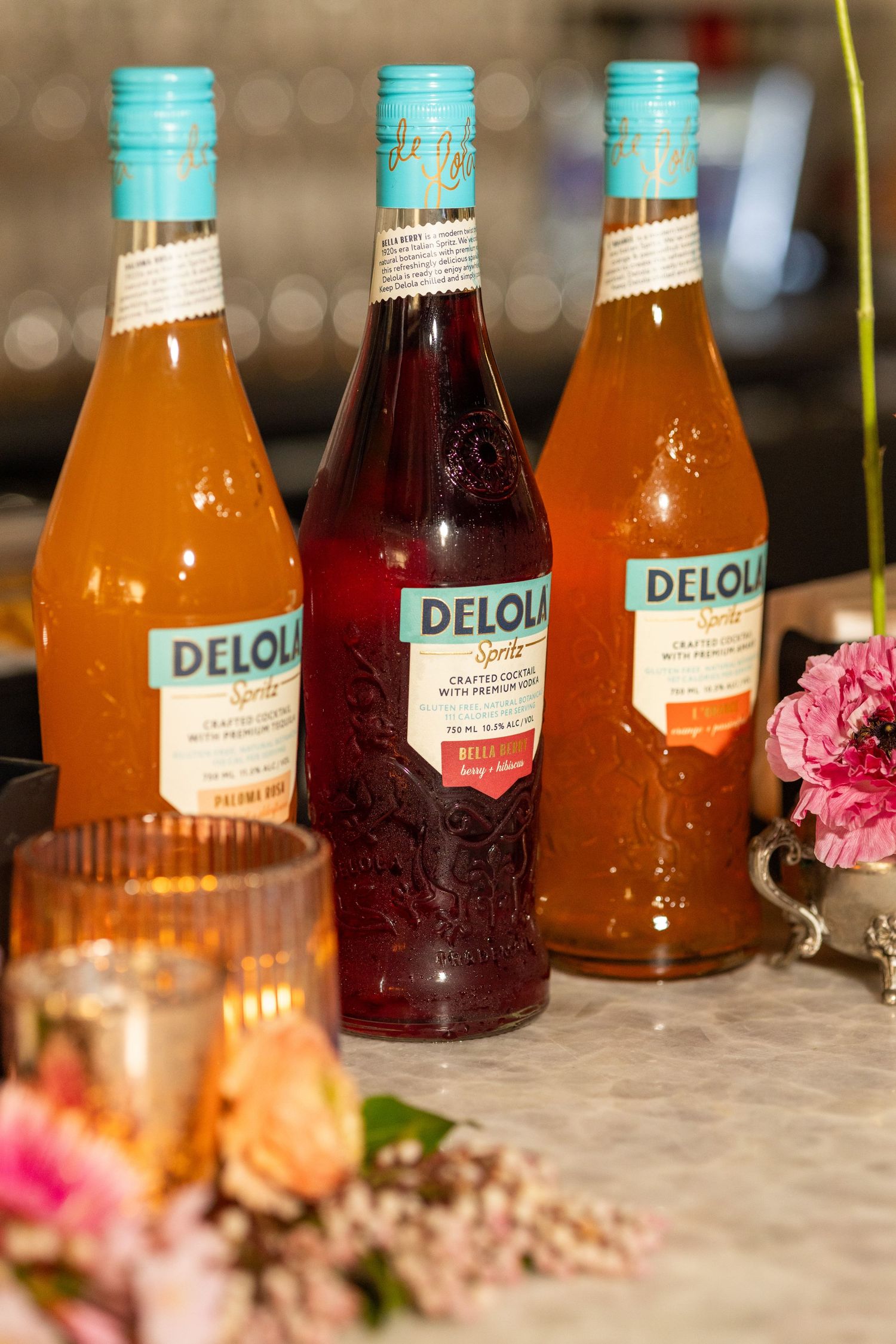 Three bottles of Delou brand beverages displayed with pink flowers in the foreground.