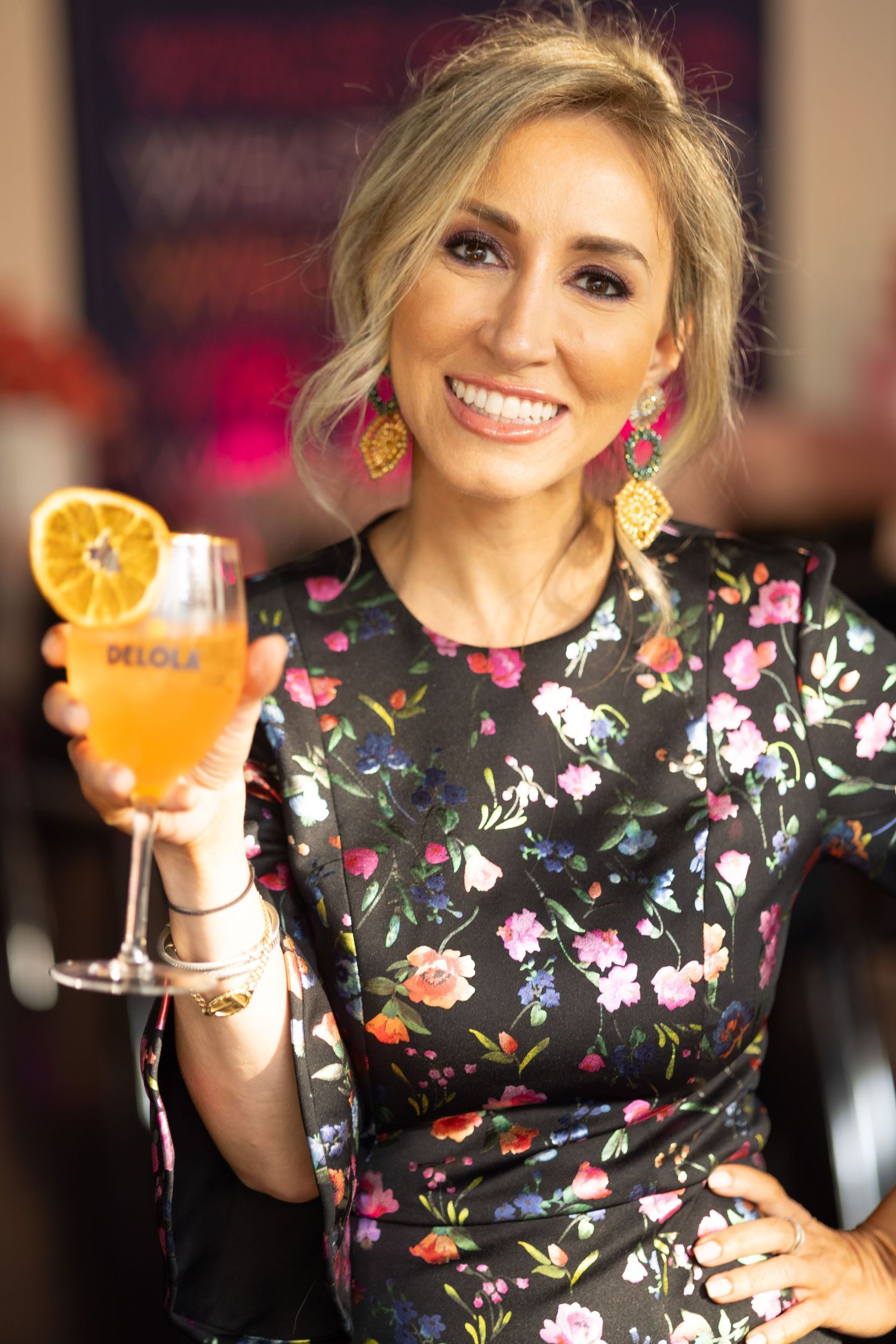 A floral dress-clad woman holds up an orange cocktail while smiling at a social event.