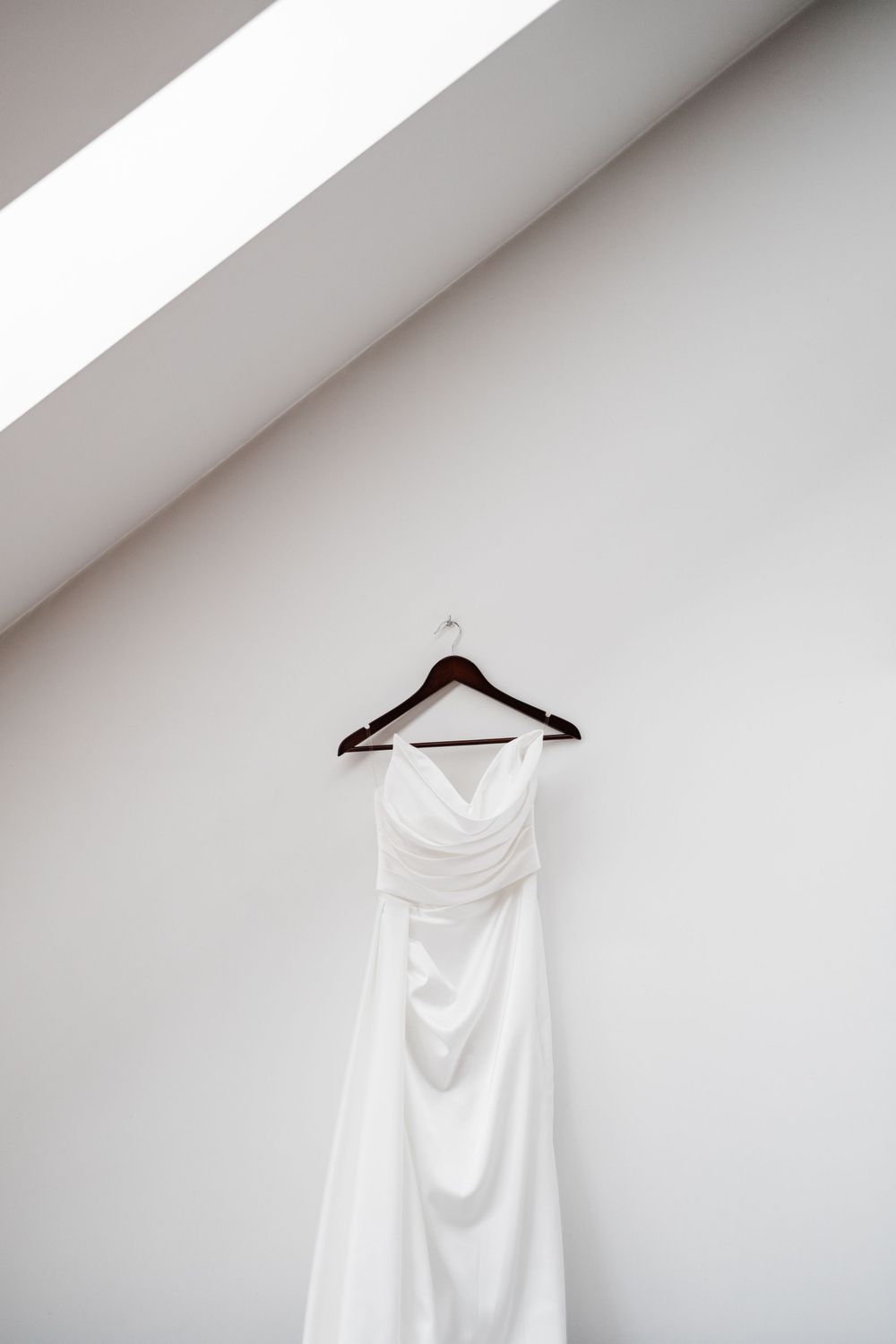 A white wedding dress hangs on wooden hanger against minimalist white wall.