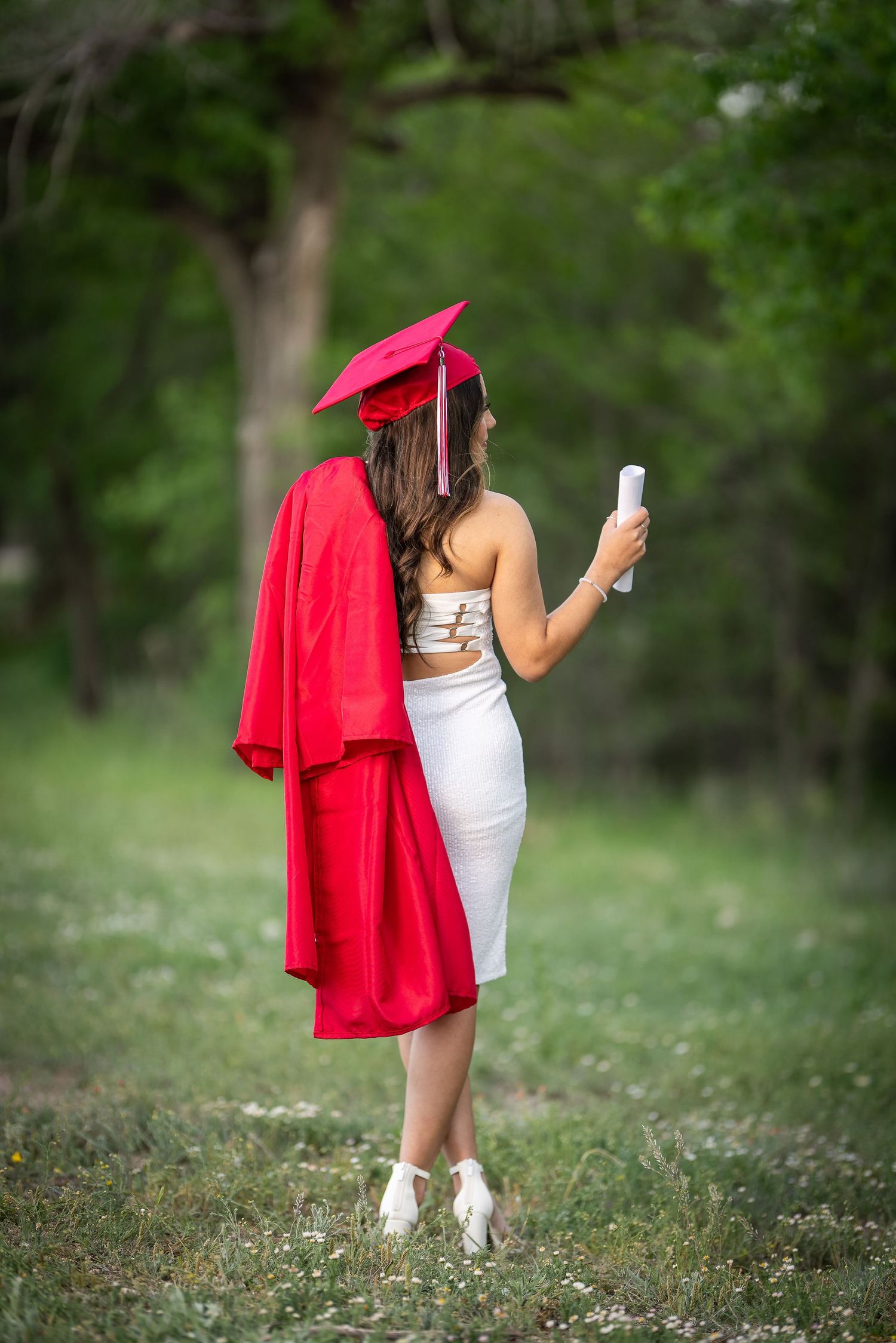 Graduate in red cap and gown holds diploma while standing in outdoor garden setting.