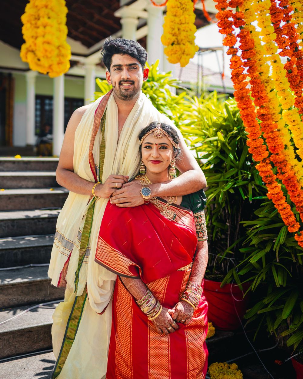 Couple in traditional South Indian wedding attire standing on steps with orange flower decorations.