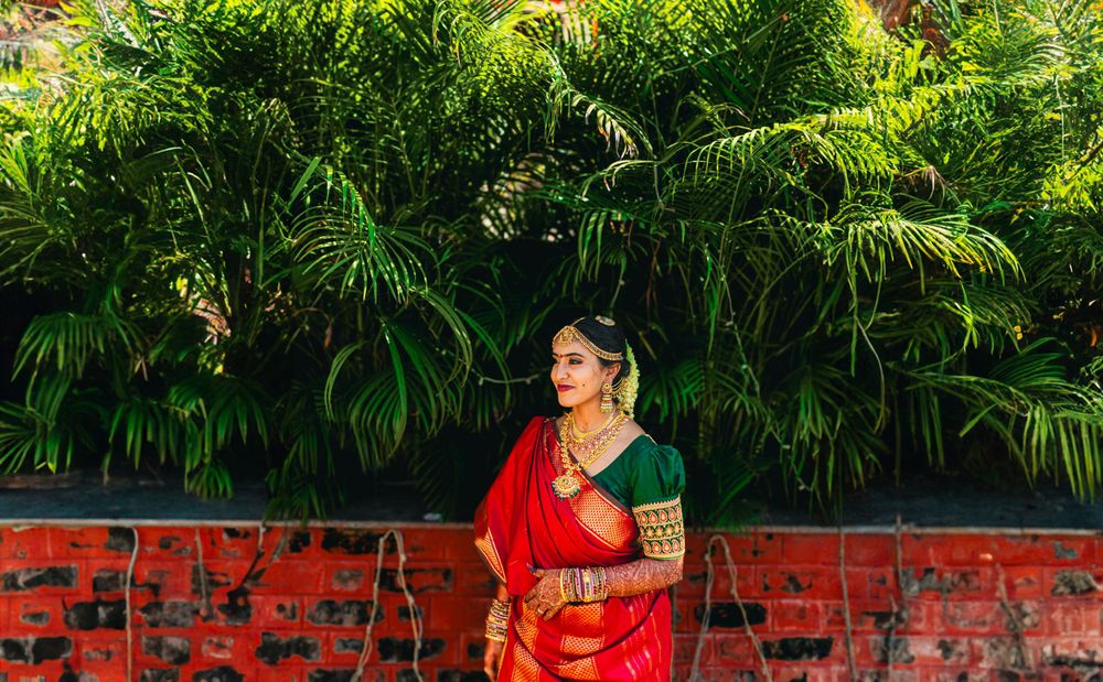 Woman in red saree posing against lush green tropical plants and brick wall.