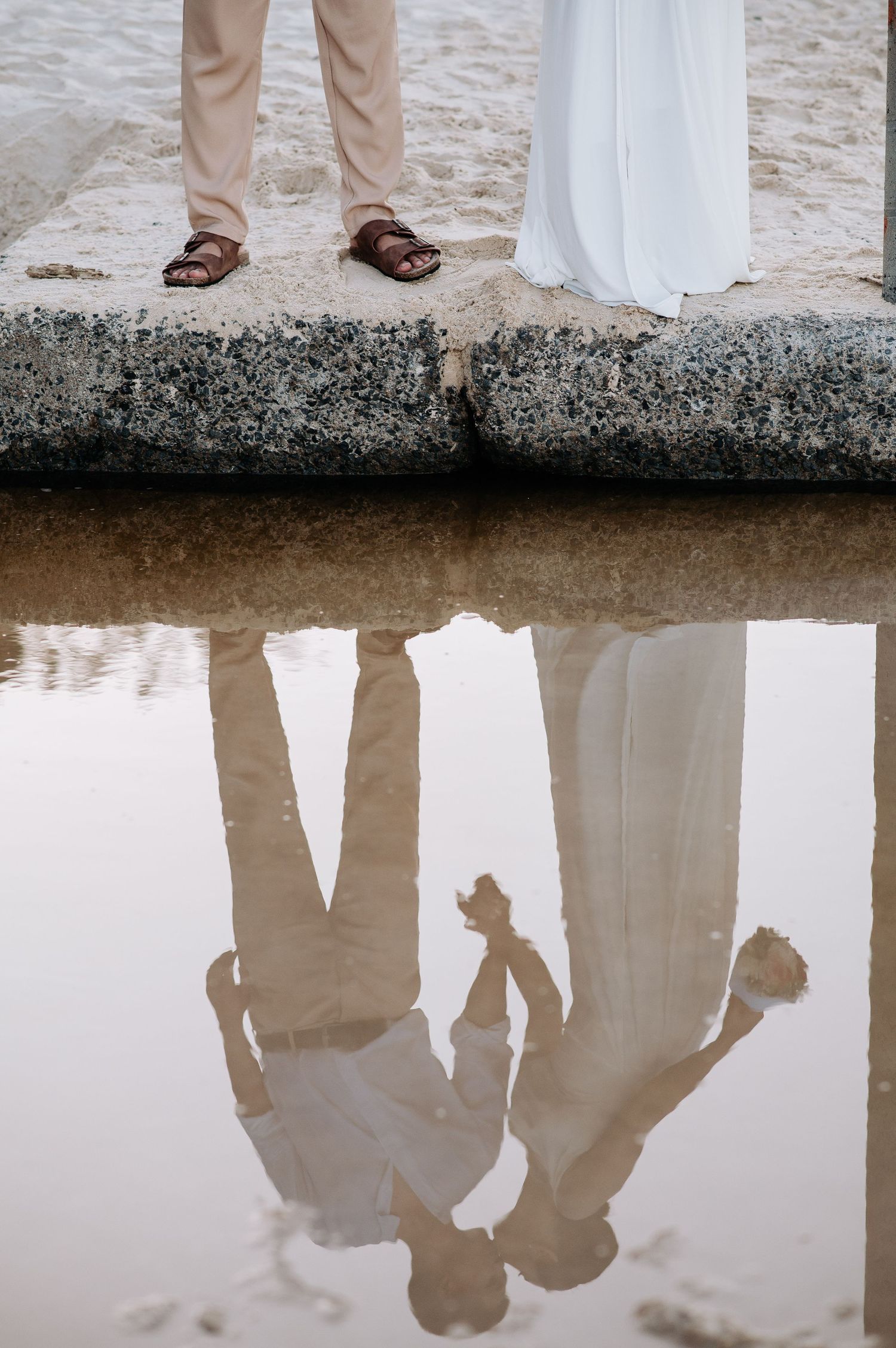Reflection of a couple in formal wear standing on concrete by the water's edge.