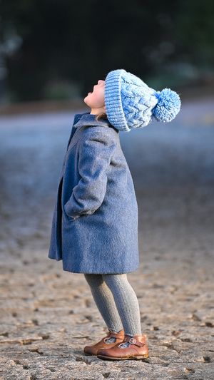 A small child wearing a pretty blue coat, gray tights, brown shoes and a blue knit hat standing under an arch.