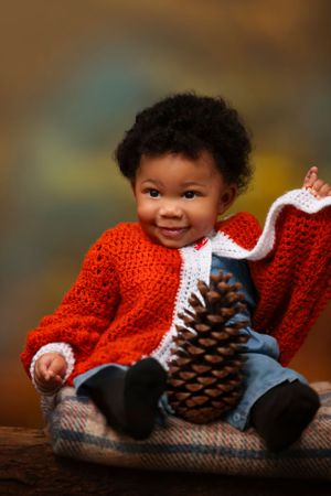 A small child wearing a red knit sweater sits on a blanket with a large pinecone, smiling warmly against a blurred background.