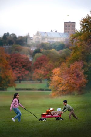 Children playing with a red wagon on a grassy hill during autumn with buildings visible in the background.