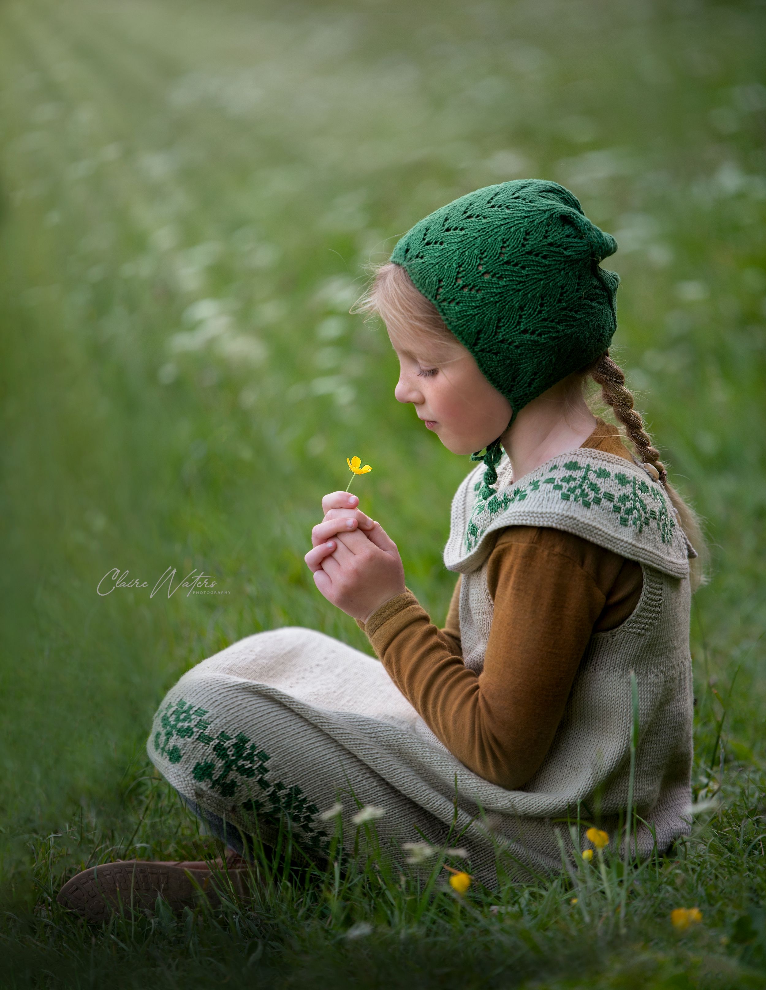 A child in a green knit hat and patterned vest sits peacefully in a meadow examining a small yellow wildflower.