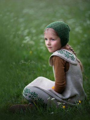 Child wearing a green knit hat and brown sweater sits thoughtfully in the grass on a soft-focus summer day.