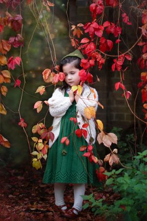 A girl  in a green knit dress stands beneath bright red autumn foliage and vines against a dark brick wall.