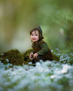 A small child in a green knit outfit sits among moss and delicate wild flowered garlic in a dreamy woodland setting.