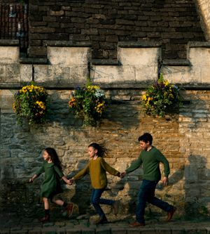 three siblings running along a Cotswolds old road with historic building behind them, playing