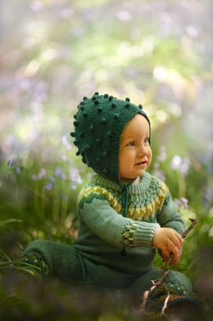 A small child in a green knitted sweater and matching bobble hat sits in a garden with soft purple flowers in the background.