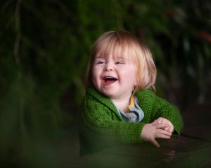 A joyful toddler wearing a green knit sweater laughs against a dark background.