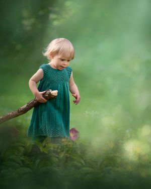 Small child in green dress holds wooden stick while exploring misty forest setting in dreamy nature scene.