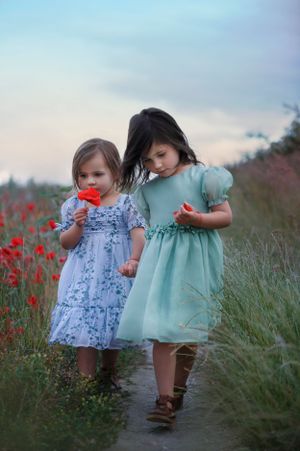 Two young children in vintage style dresses walking together through a field of red poppies at sunset.