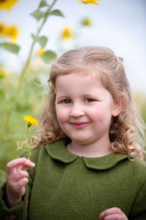 A child in a green heirloom handmade dress with a Peter Pan collar smiles while holding a yellow flower in a sunny garden.