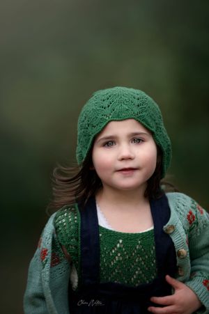 A green knitted beanie and cardigan complement navy overalls in this soft focus outdoor portrait.