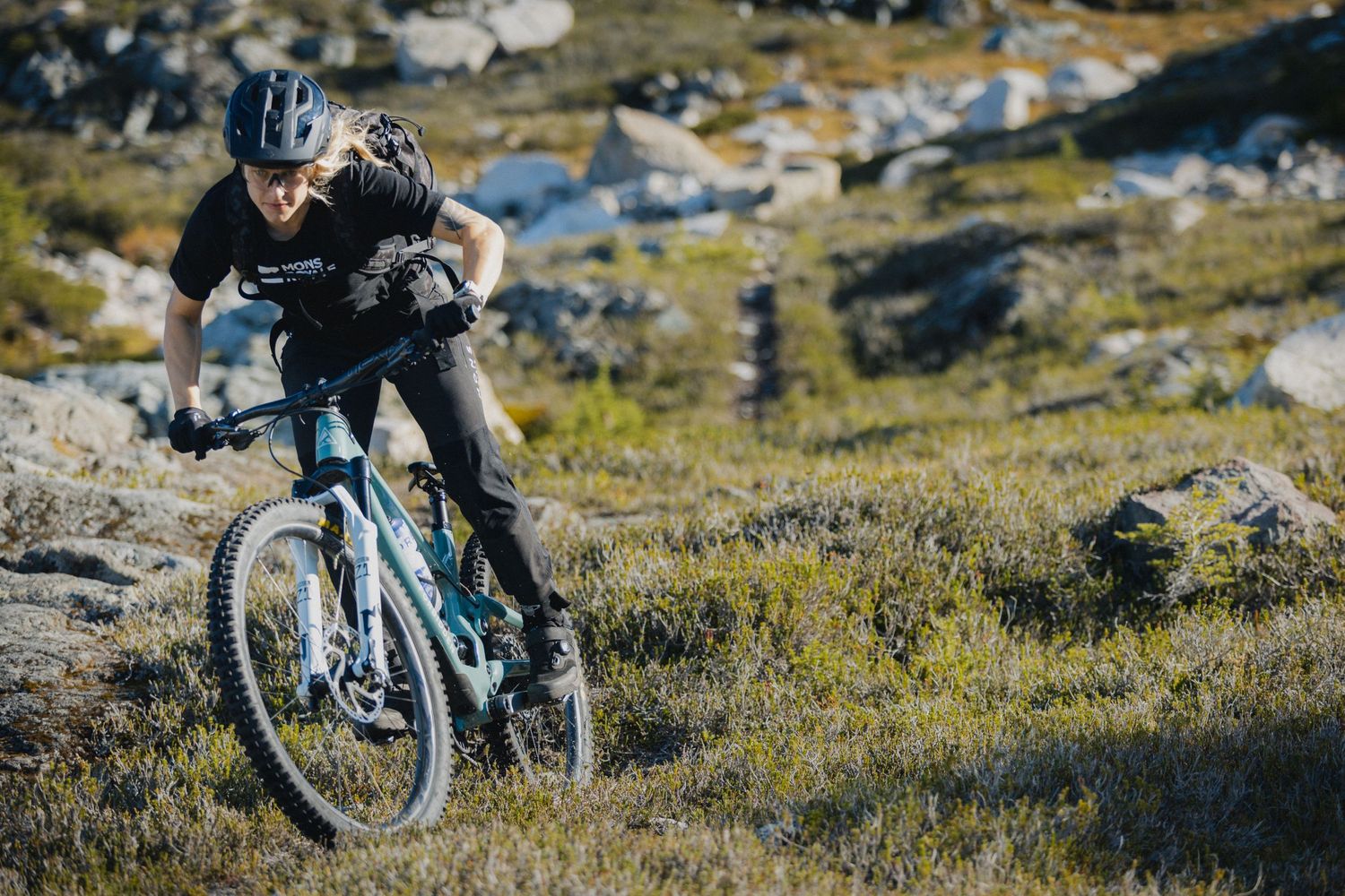 Mountain biker rides rocky alpine trail with mountain landscape in background.