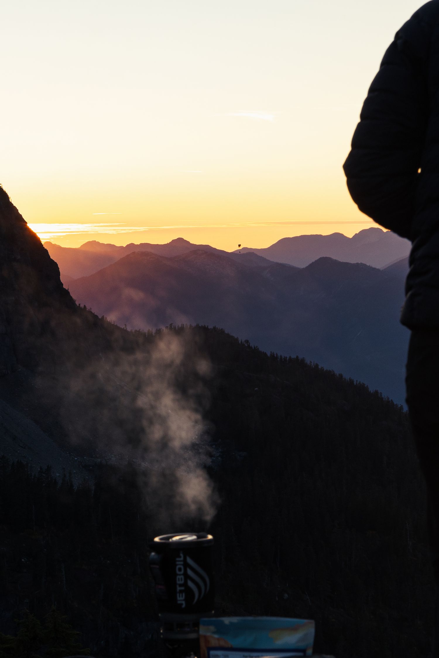 Misty mountain sunset with campfire smoke rising in silhouetted rocky landscape.