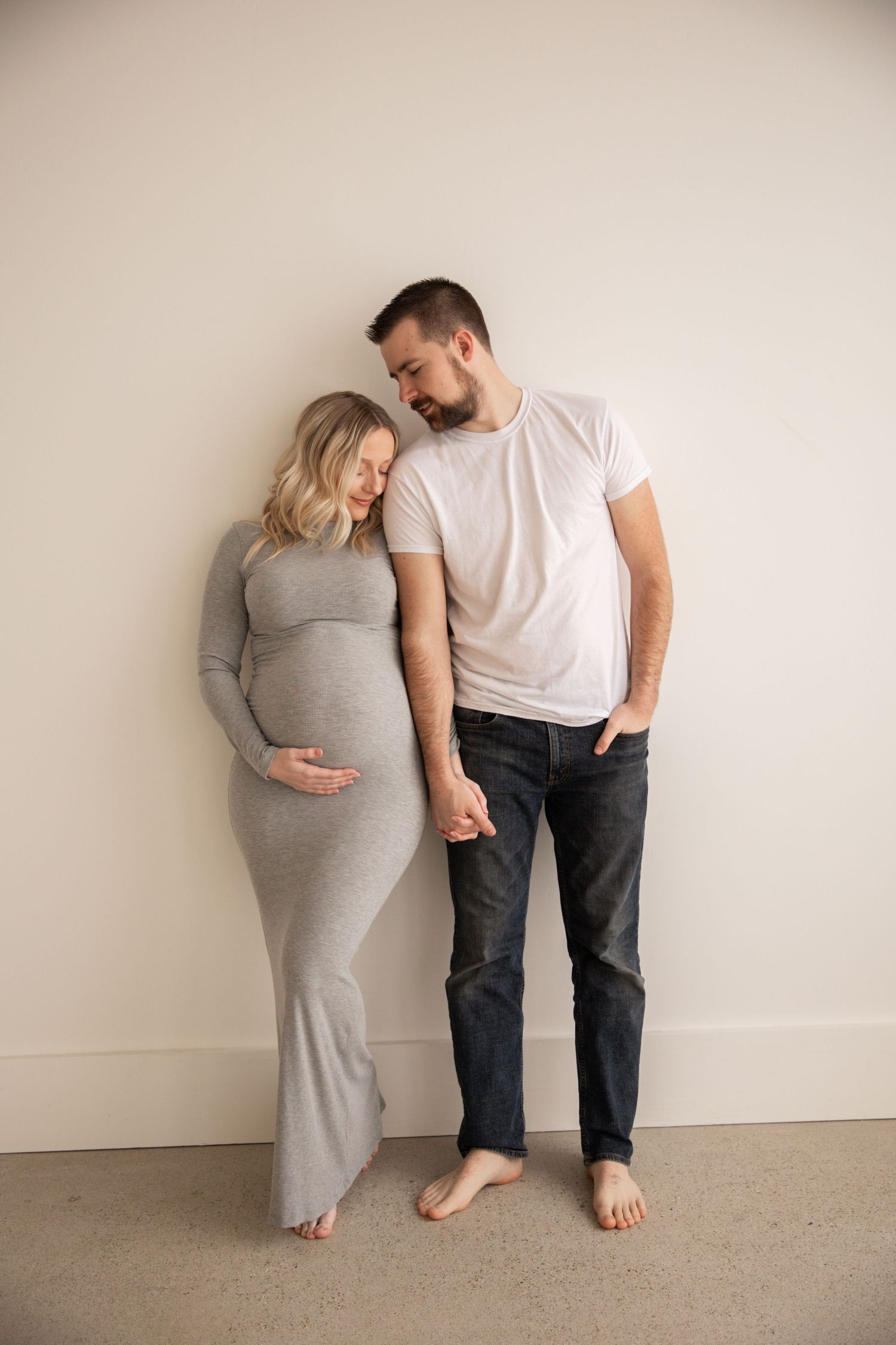 Intimate maternity photo of couple in casual clothing against neutral wall.
