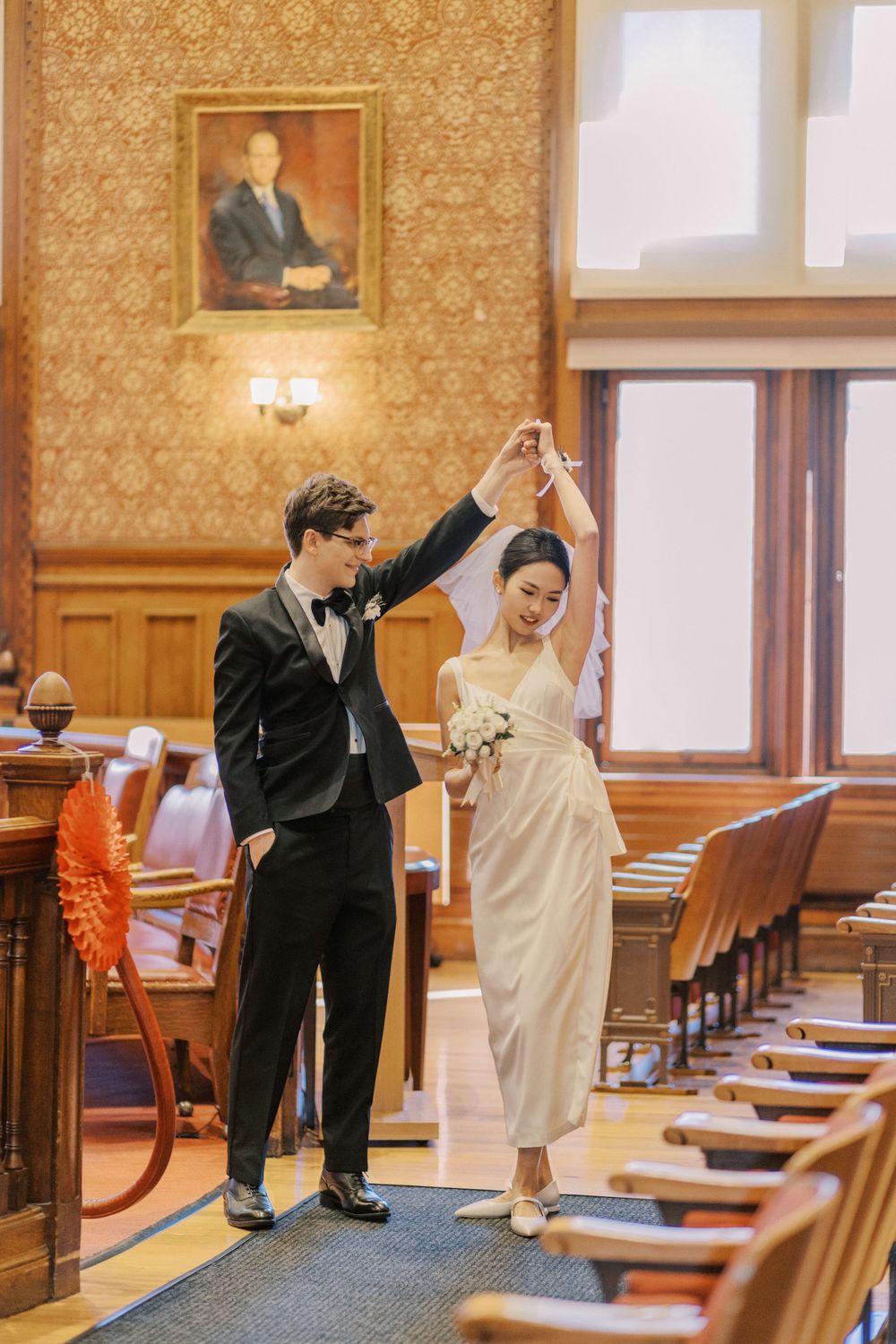 A bride and groom dance playfully in a wood-paneled hall with orange chairs.