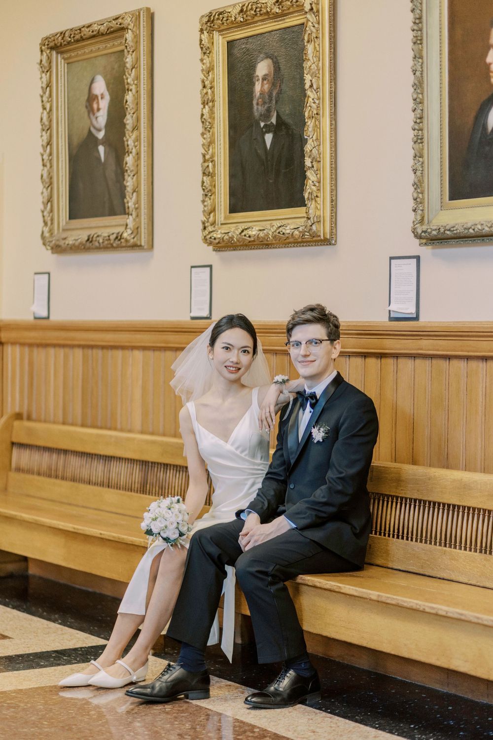 Newlyweds sit on wooden bench beneath gallery of vintage portraits in elegant hall.