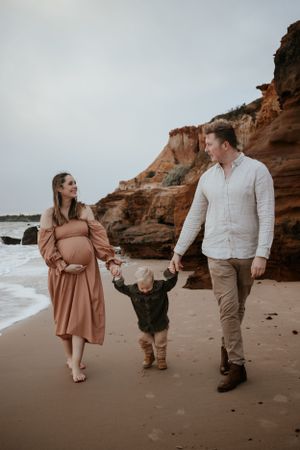 Pregnant mum and dad walking along the beach holding hands with their son.