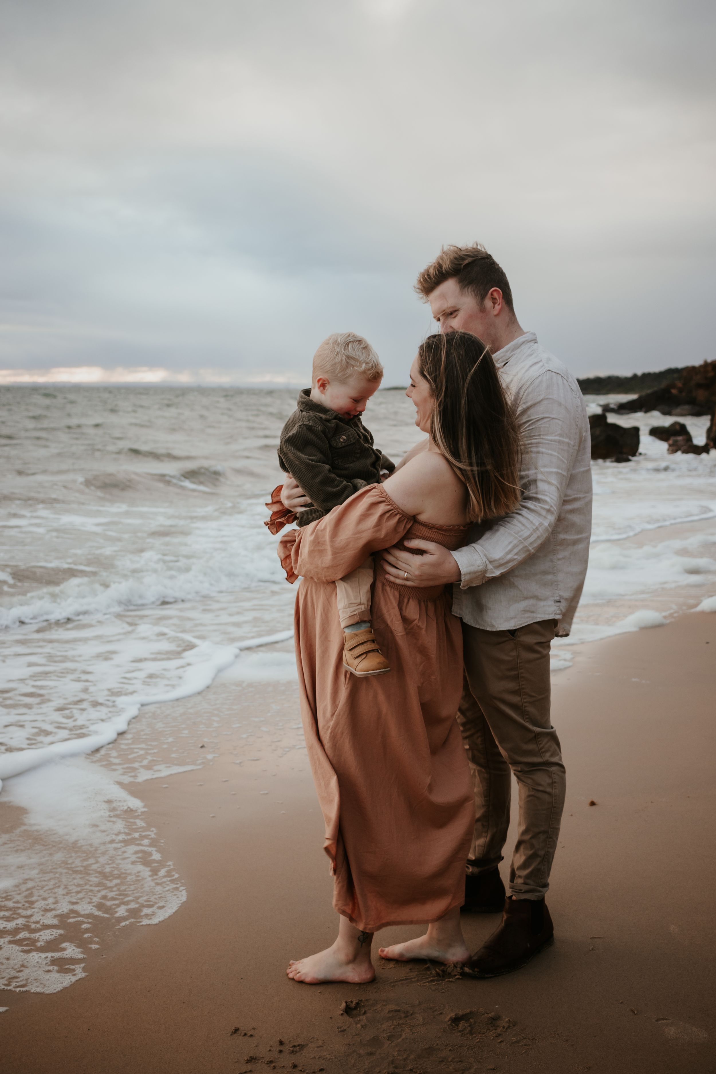 Pregnant mum and dad hugging their son at the beach.