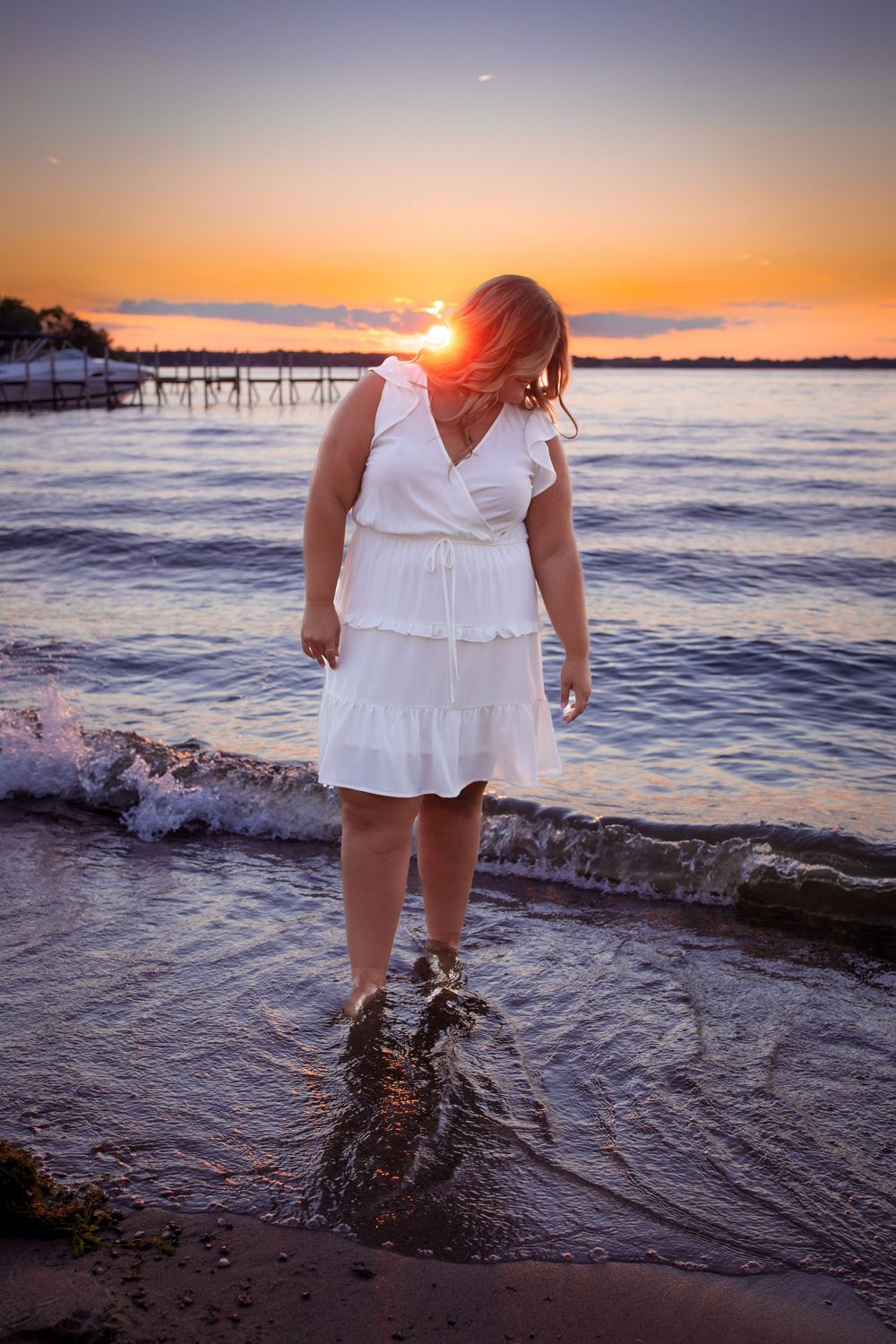 Senior Session of a girl in a white dress standing in shallow water with West Lake Okoboji and a colorful sunset in the background.