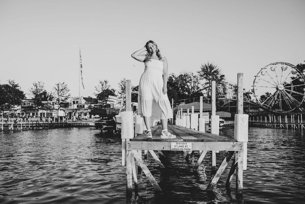 Black and white Senior portrait of a girl in a white dress standing on dock with Arnold's Park Amusement Park in the background.